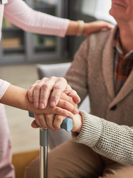 Close-up of young caregiver holding hands with elderly man and talking to him, she caring about him at home