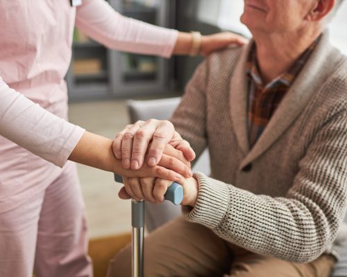 Close-up of young caregiver holding hands with elderly man and talking to him, she caring about him at home